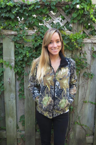 Woman wearing a camouflage jacket standing in front of a wooden fence with greenery.