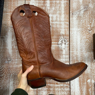 A person's hand holding a tan-colored leather cowboy boot with a round toe and a heel.